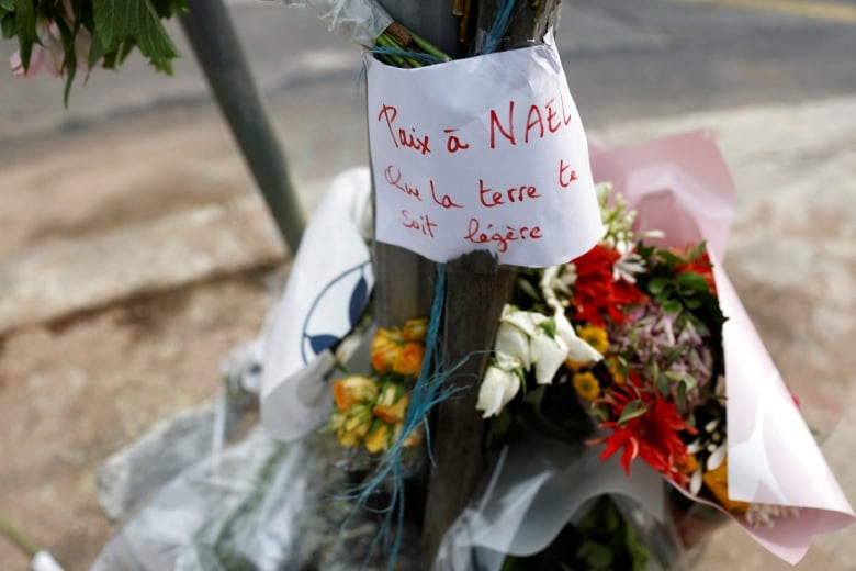 Bouquests of flowers lean against a pole. A message above them reads: "Peace to Nael, may the earth be light to you."