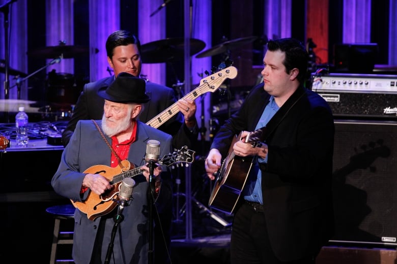 Three musicians on stage. Two younger men play guitars, while and older man with a long white beard plucks a mandolin.