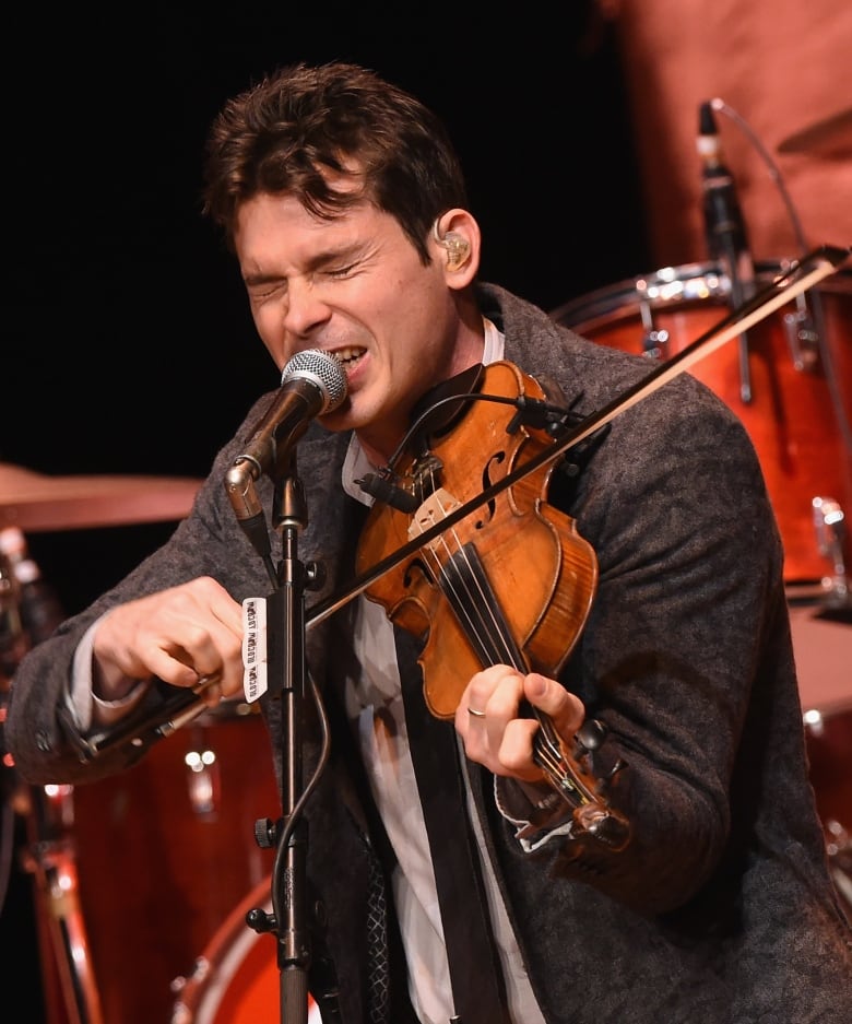 A man with short brown hair squeezes his eyes shut as he sings and plays the fiddle.