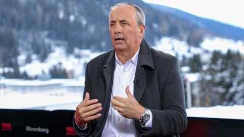 A middle-aged man in a suit speaks to a camera with a winter landscape behind him