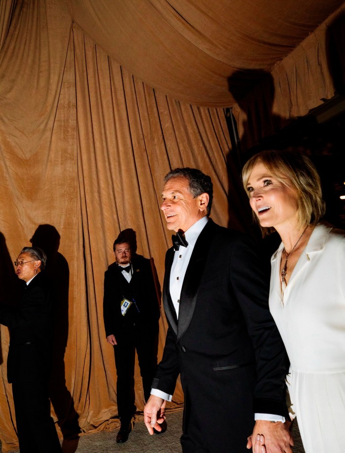 A man in a tuxedo and a woman in a white outfit, both smiling, walk hand in hand