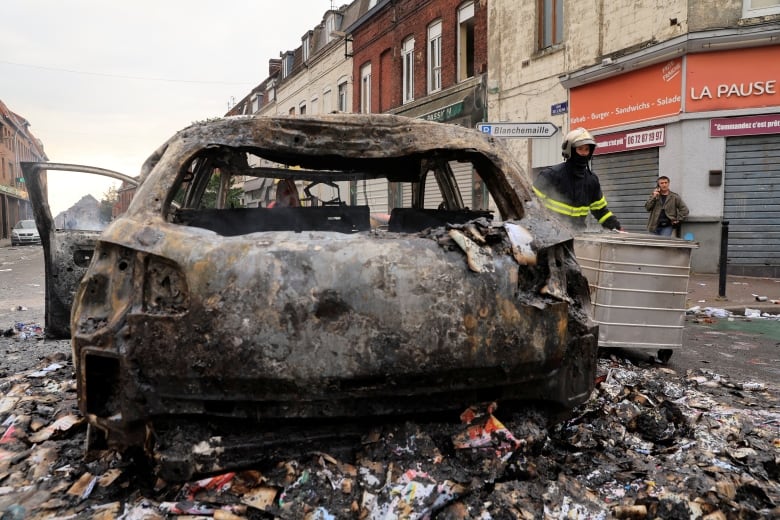 The burned-out hull of a car sits in a street. 