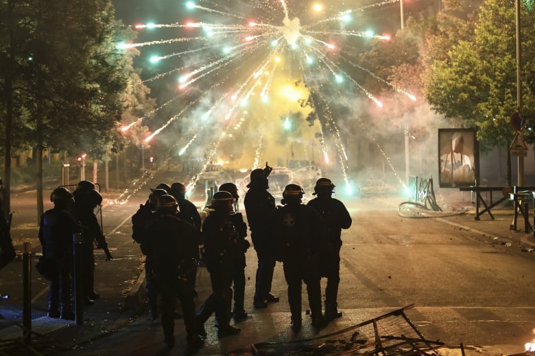 Ten or so people, at least some wearing visored helmets, stand in a street at night. Arcs of red and green light radiate outward from an apparent firework detonation.