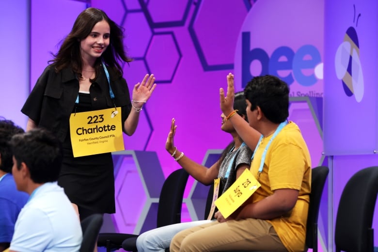 A girl smiles and gives a high-five to two seated people. She wears a yellow sign that says "Charlotte". The wall behind them says "bee".