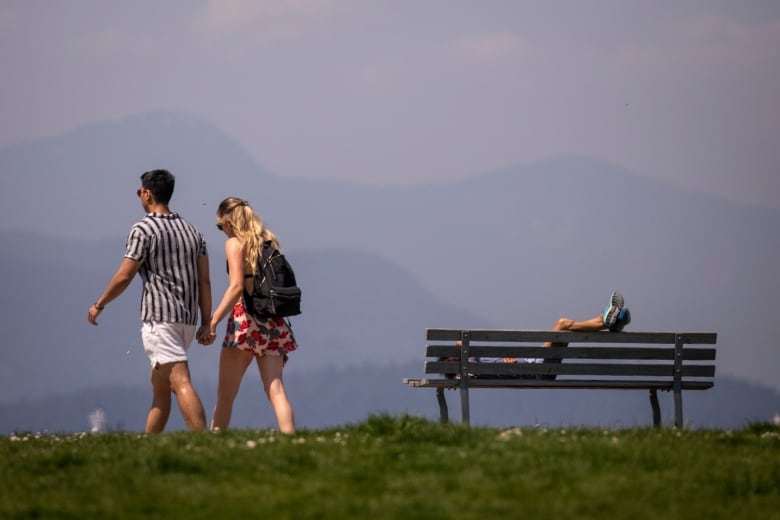 One person lies on a park bench while two people walk by amid hazy, smoky air seen in the mountainous landscape. 