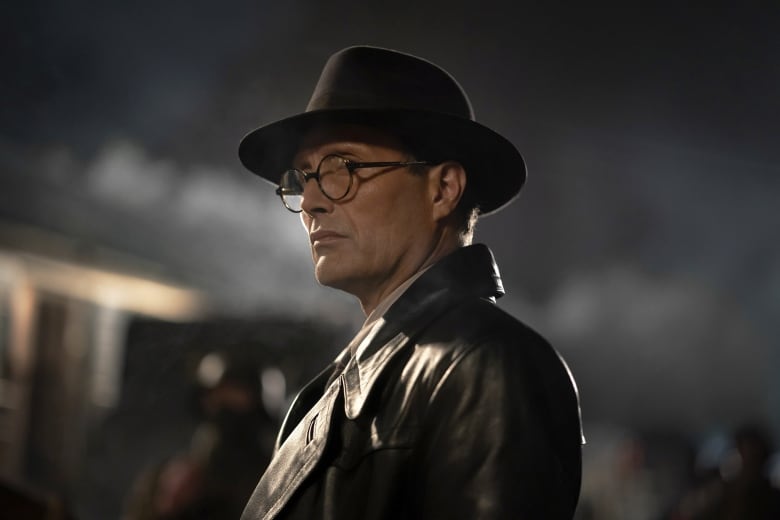 A man in round eyeglasses, a leather jacket and a fedora looks offscreen. 