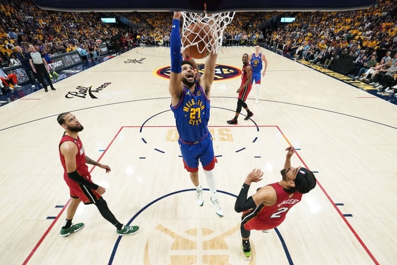 A male basketball yells while hanging from the rim with both hands as the ball falls through the net and opposing players watch from below.