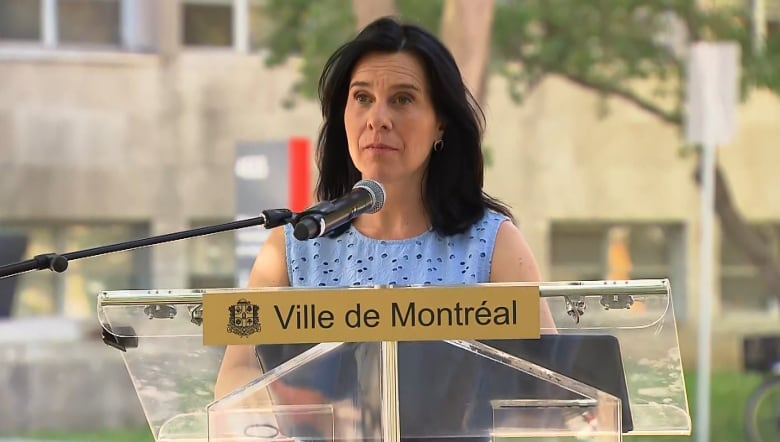 Montreal Mayor Valérie Plante stands at a clear city podium at an outdoor news conference on a hot sunny day, wearing a light blue top.