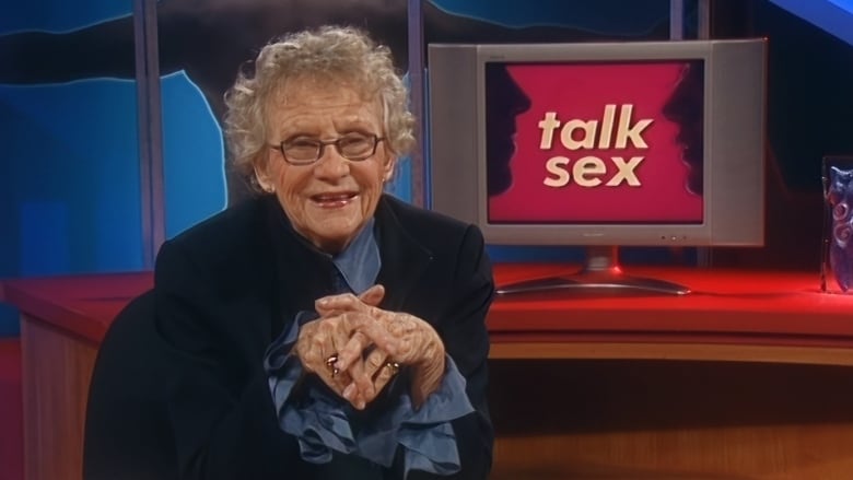 An elderly woman sits at a desk with a pen and pad of paper. A screen behind her showing the words "talk sex."