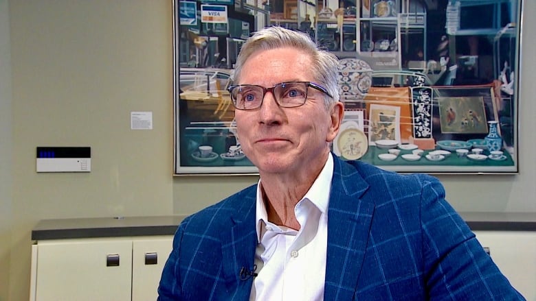 A man in a suit is sitting in a boardroom with a large painting hanging on the wall in the background.