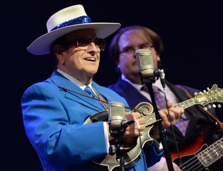 A man in a bright blue suit, a matching sequin hat and big sunglasses smiles and plucks a banjo.