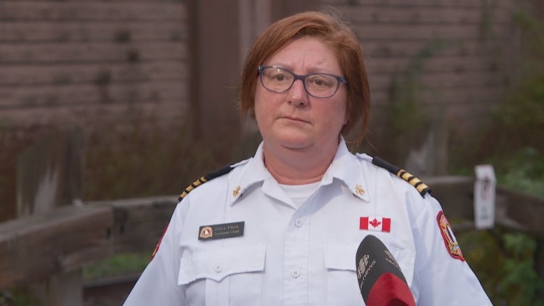 A woman with short red hair and glasses, wearing a white tunic uniform, stands in front of a CBC microphone.