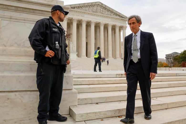 A man in a suit is shown walking by a police officer near the steps of the U.S. Supreme Court.