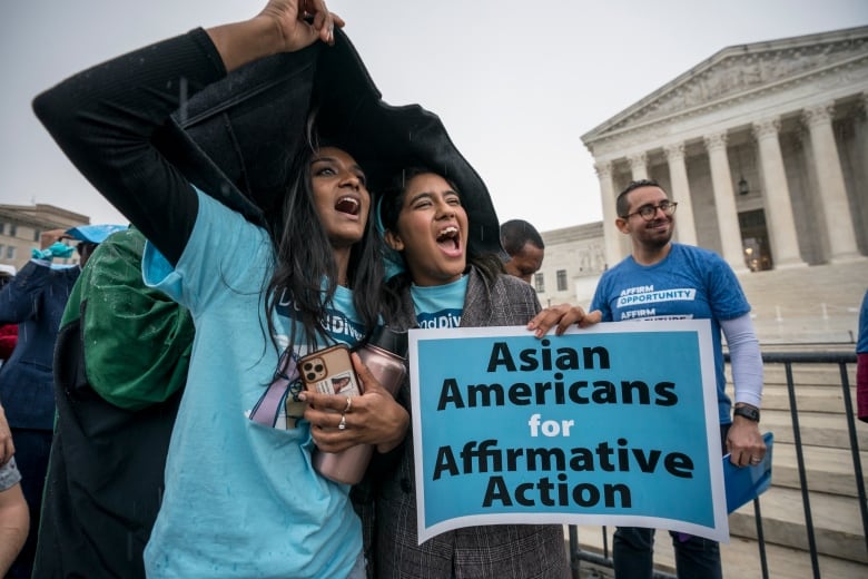 Two young women cover their heads with a jacket from the rain while holding a sign reading 'Asian Americans for affirmative action' below the steps of the top court in Washington.