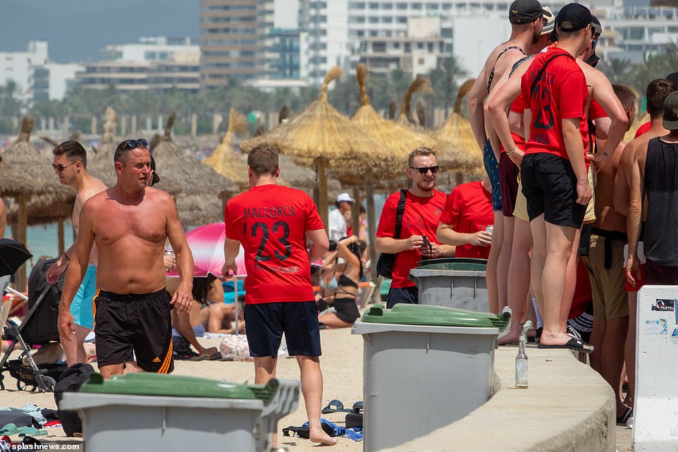 A group in red drink beers as they stand beside the beach