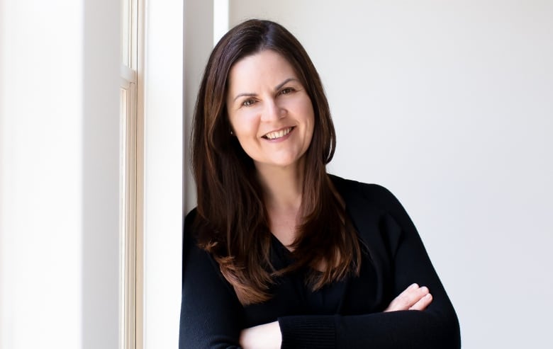 A woman leans against a wall, arms folded.