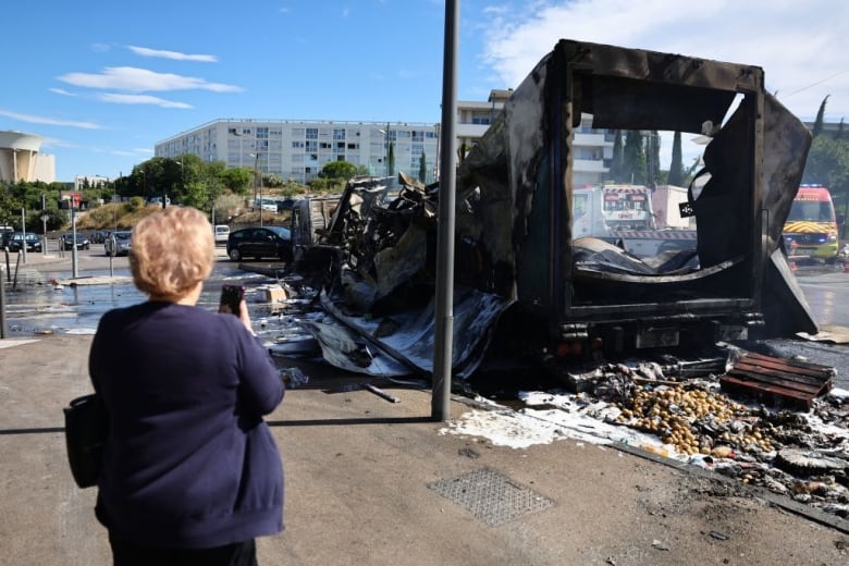 A woman takes a photo of a burned-out bus.