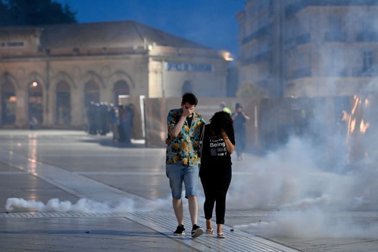 Two people in civilian clothing walk among clouds of tear gas, hands to their faces.