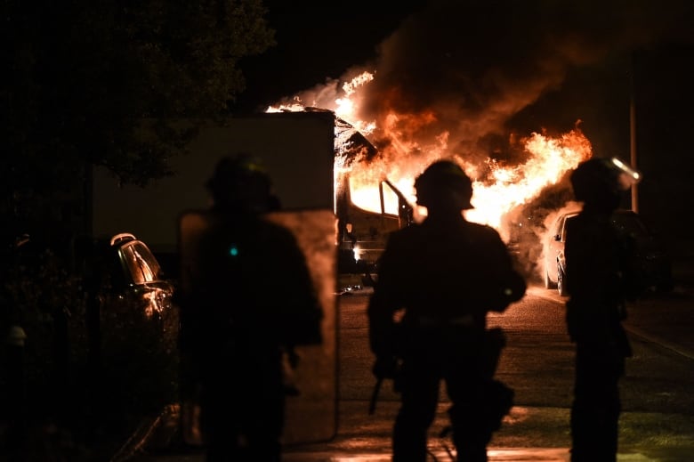 A few silhouetted figures stand before a fire on a street a night.