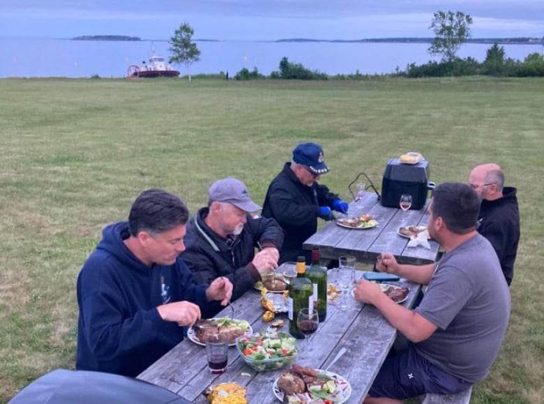 Five men sit at a picnic table for supper at dusk, with the hovercraft docked behind them.