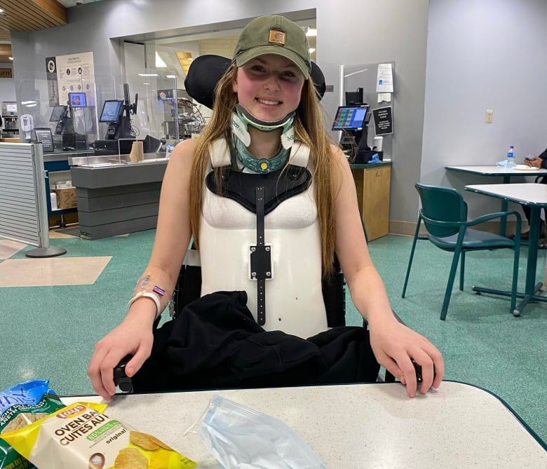 A young woman is sitting at a table in a hospital cafeteria. She wears a full torso cast and neck brace. She has orange-ish hair and is wearing a green ball cap. She's smiling for the photo. 