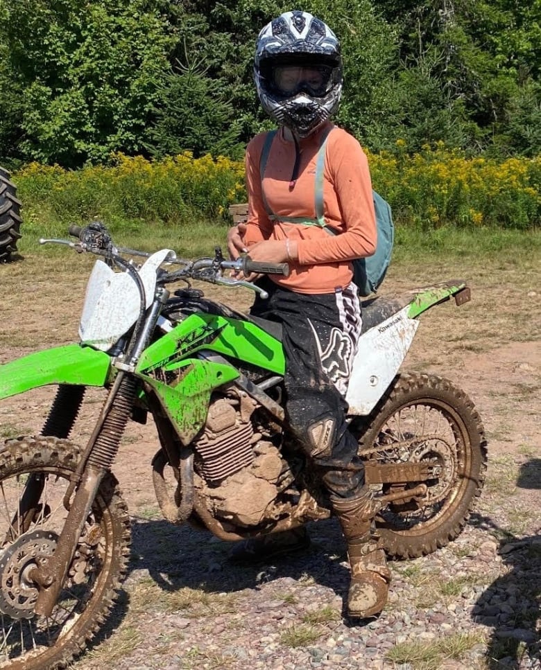 A young woman sits on a lime green dirt bike. It's covered in mud. She's wearing a full helmet and an orange shirt. There are bright green trees in the background. 