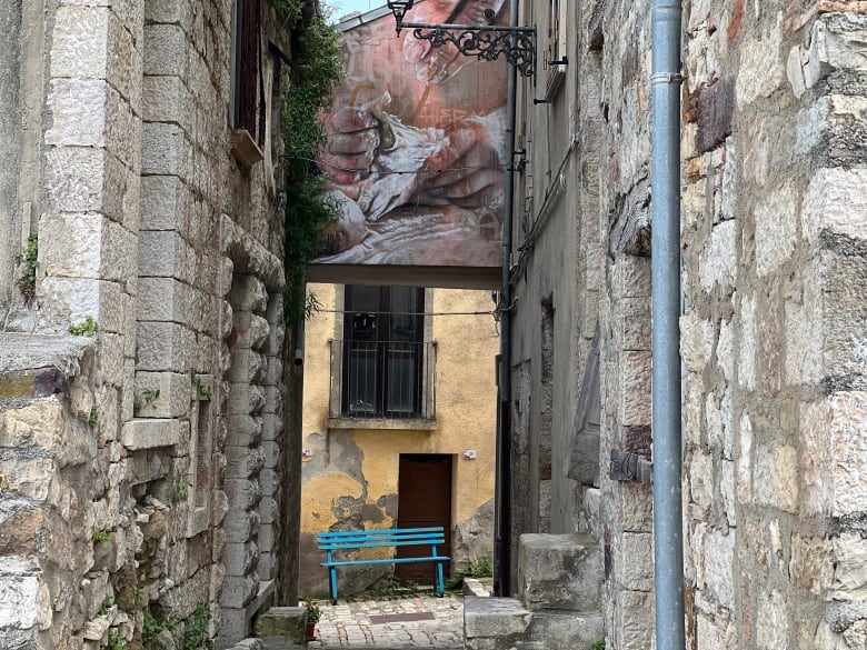 A mural of hands is shown on a stone arch of a narrow alley. At the end of the alley sits a bright blue bench.