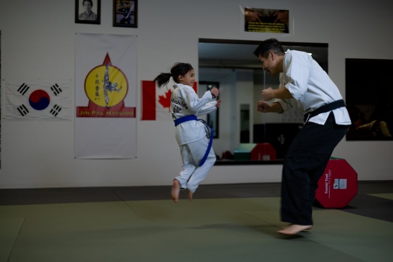 A young girl, captured mid-leap, spares with her dad in a dojang. Both wear white gis. She has a blue belt and him, a black one.