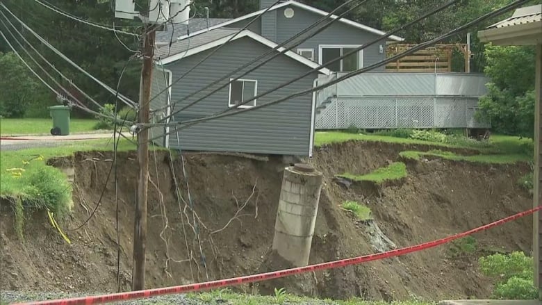 A shed with grey siding is very close to the edge of a hole caused by a landslide, with a corner of it over the edge. Nearby house with matching grey siding sits only slightly back from the edge.