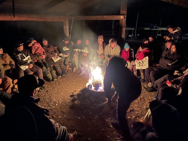 A group of people sit around a fire in the dark.