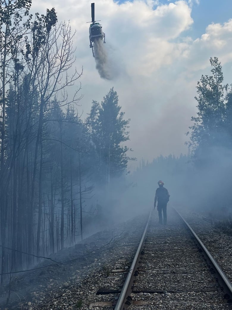 A firefighter, shrouded in smoke, looks on as a helicopter sprays water above him.