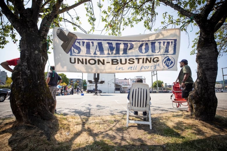 A large sign strung up between two trees reads "Stamp out. Union-busting in all ports." 