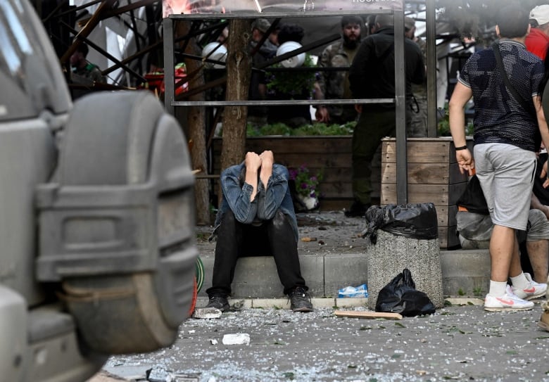 A man sits on a curb, head buried in his hands, debris strewn around him.