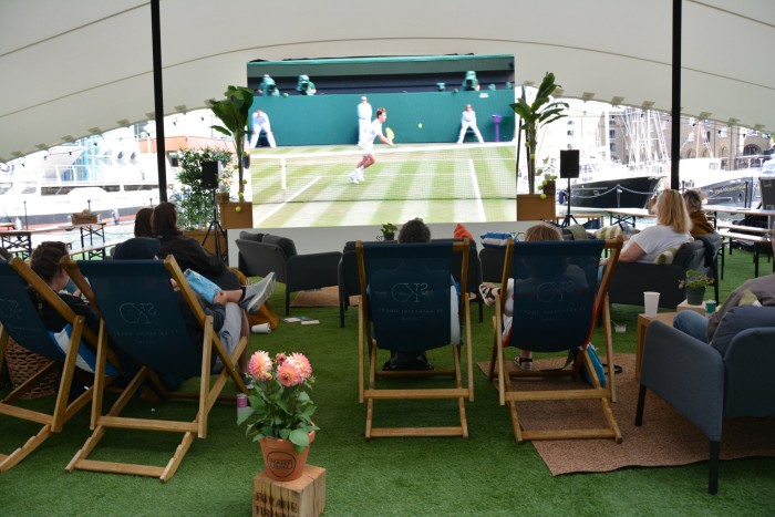 People in deck chairs watching Wimbledon on a big screen on a pontoon floating in London’s St Katharine Docks