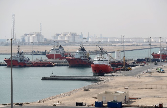 Three ships are docked at a water inlet with industrial facilities in the distance