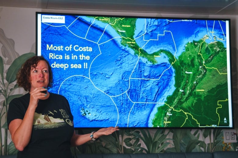 A woman speaks into a microphone in front of a screen with a map showing parts of South America and the Pacific ocean, and the words "Most of Costa Rica is in the deep sea!!"