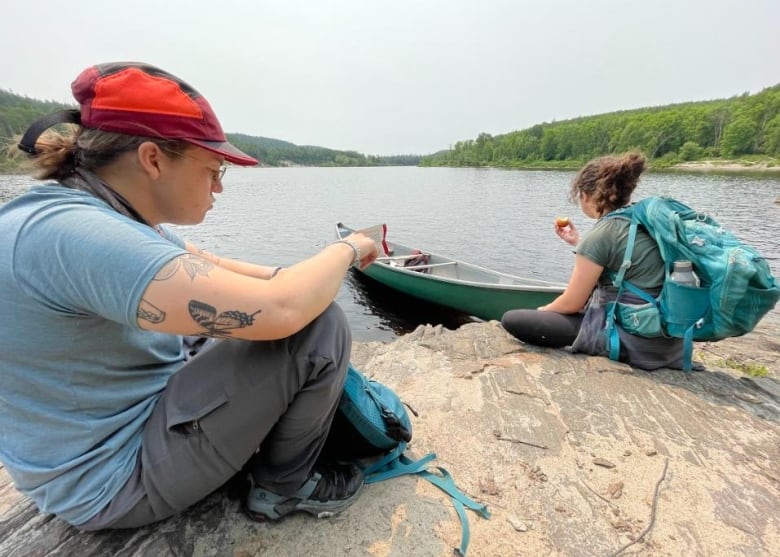 Two people sit on a sandy beach with a canoe.