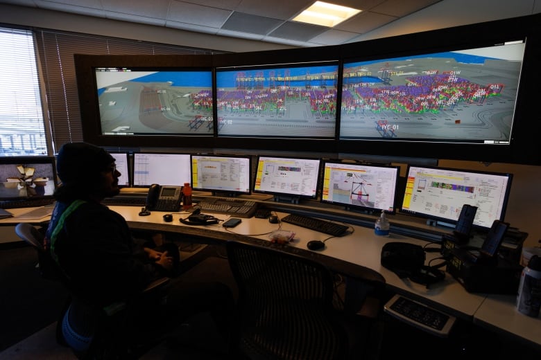 A man sits in an office in front of three large screens above a row of six computer monitors.