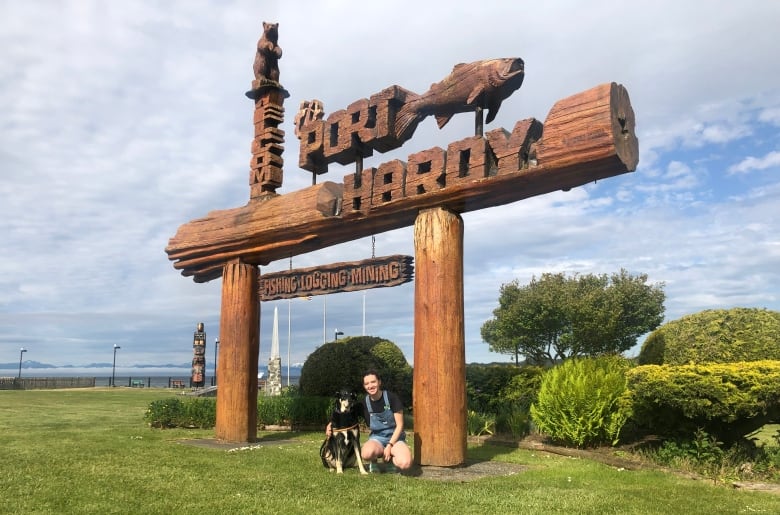 A woman kneels beside a dog under a large, wooden sign for Port Hardy. 