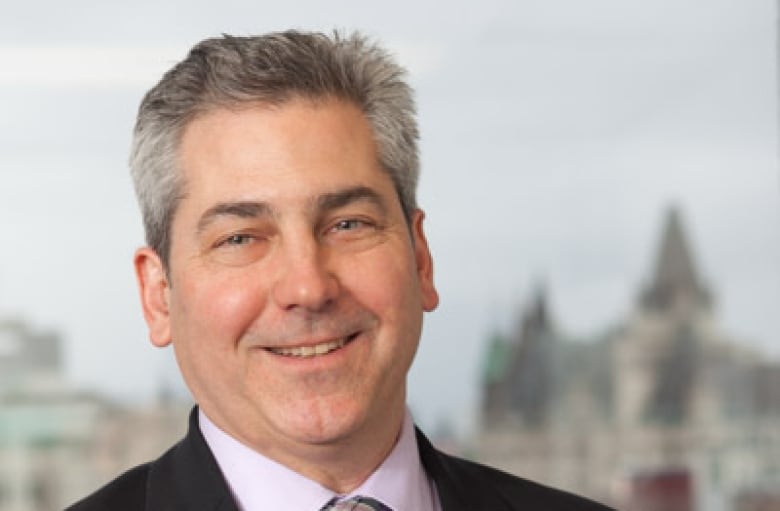 Close up headshot of John Lawford, with blurred buildings in the background. 