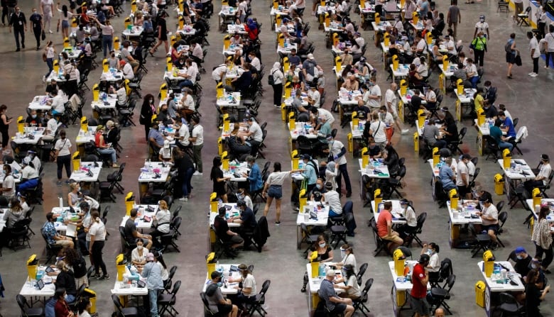 People sit at tables in an arena waiting to be vaccinated.