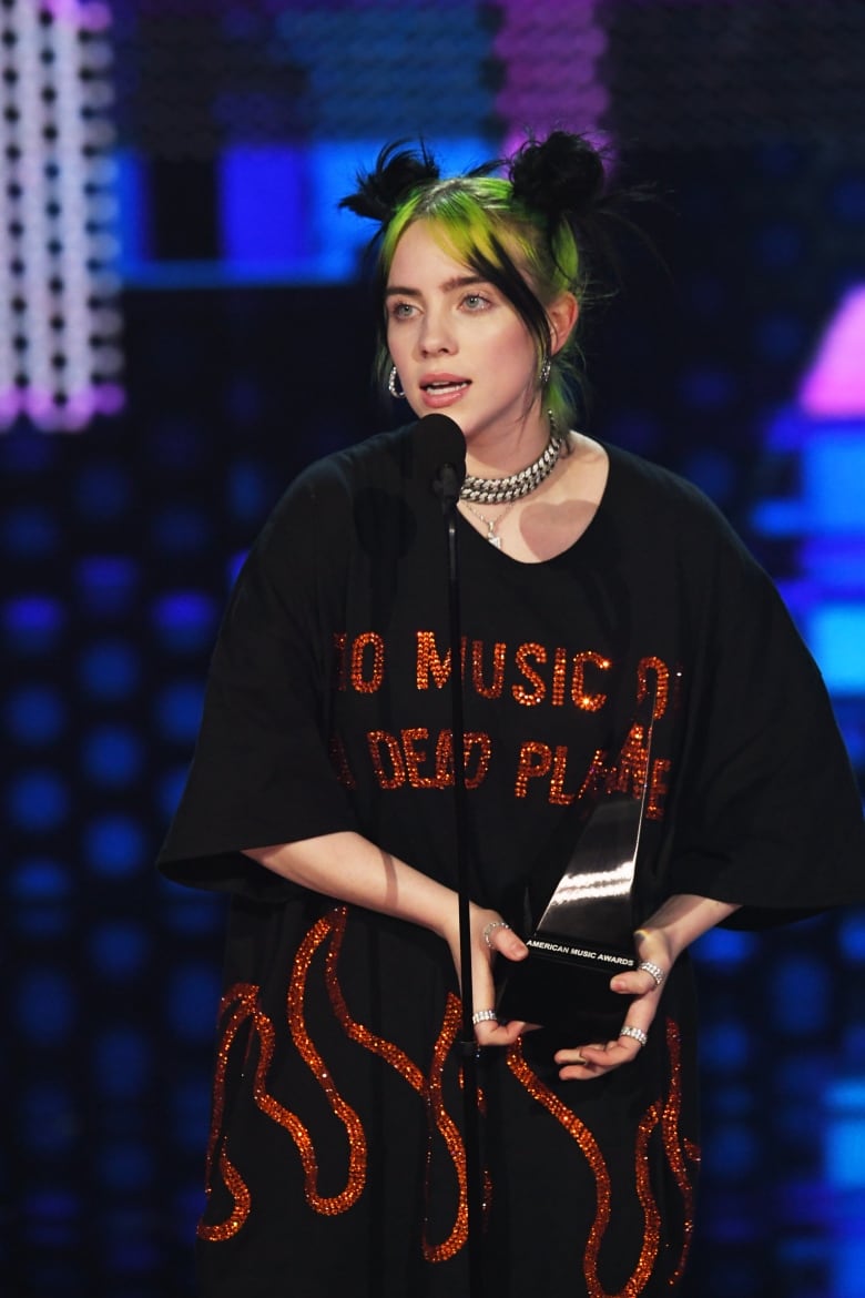 Billie Eilish, with her green and black hair in two buns, wears a t-shirt that says "no music on a dead planet."