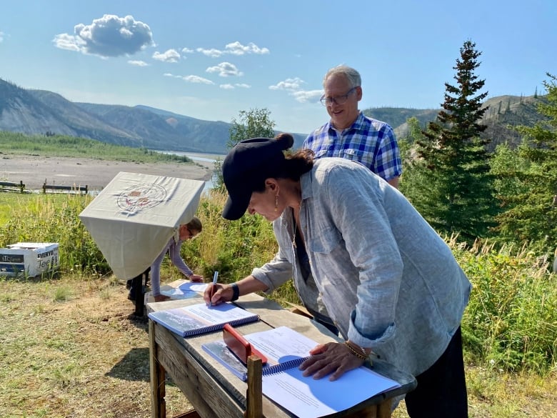 A woman stands outside a table signing a document as a smiling man looks on. Trees and mountains and a river are in the background.