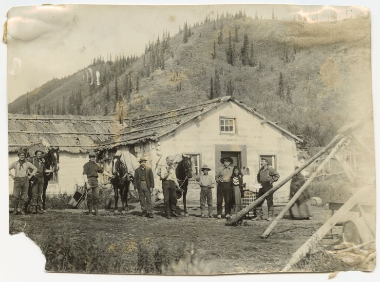 An old sepia photo of a group of men and horses standing in front of a wooden building.