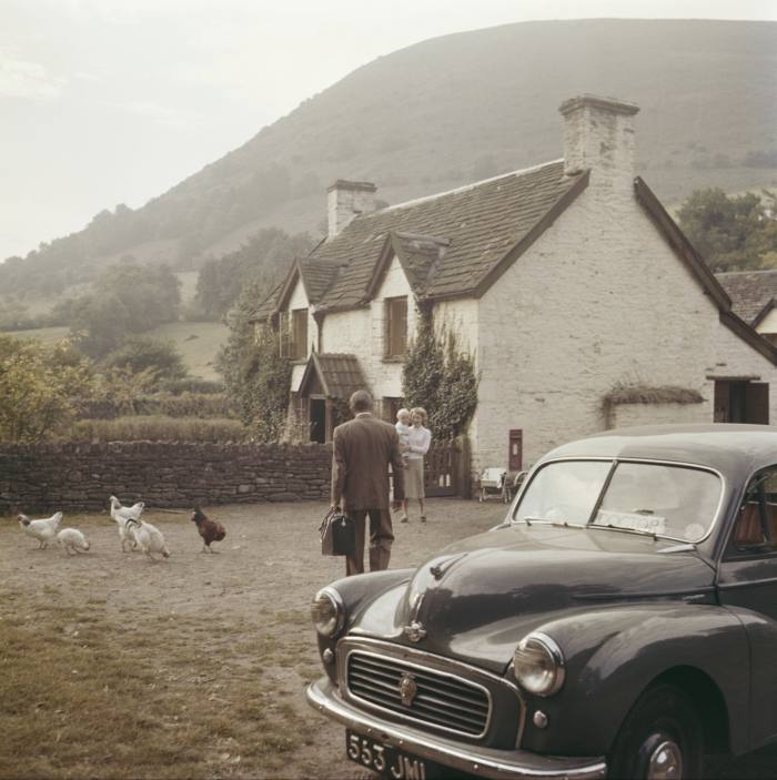 A man carrying a doctor’s bag walks from his parked car towards a farmhouse where there are chickens