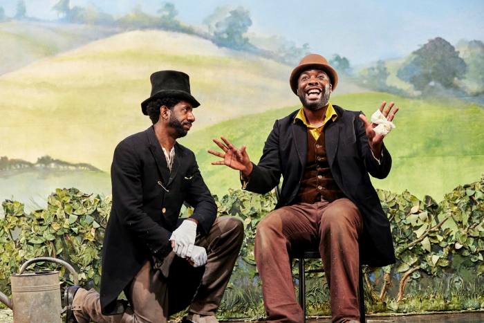 Two black men in archaic clothing and hats sit in front of a stage backdrop depicting open countryside