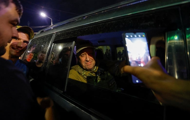 A person looks out a vehicle window as a cellphone held in front of him records the scene and other people look on.