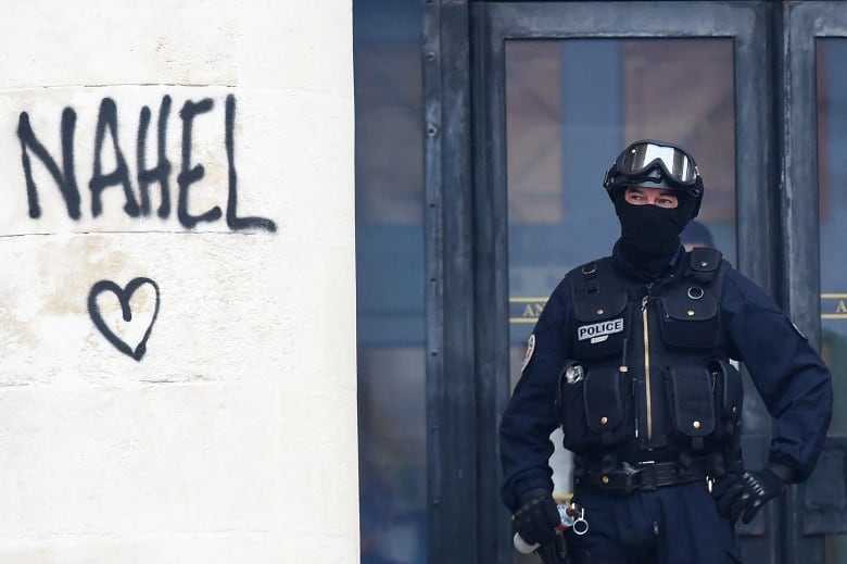 A police officer only showing his eyes stands next to graffiti that says "Nahel".