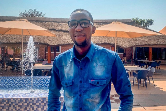 An African man in a blue denim shirt and glasses stands outside a cafe