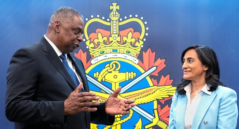 A man and a woman speak in front of a background with a crest.